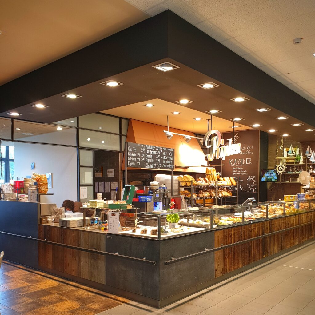 Eine moderne Bäckerei mit Glasvitrinen voller Gebäck und Brot, hölzernen Akzenten, einer Speisekarte mit Kreidetafeln und einer warmen, einladenden Atmosphäre. Ein Mitarbeiter steht hinter dem Tresen.