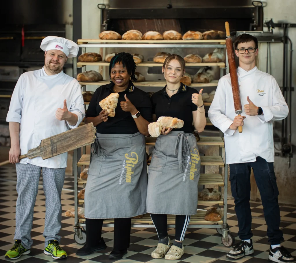 Vier Bäcker stehen lächelnd in einer Bäckerei und zeigen die Daumen nach oben. Zwei Frauen in Schürzen halten Brot, während zwei Männer mit Kochmützen ein Holzpaddel und ein Nudelholz halten. Hinter ihnen stapeln sich frische Brote auf Regalen.