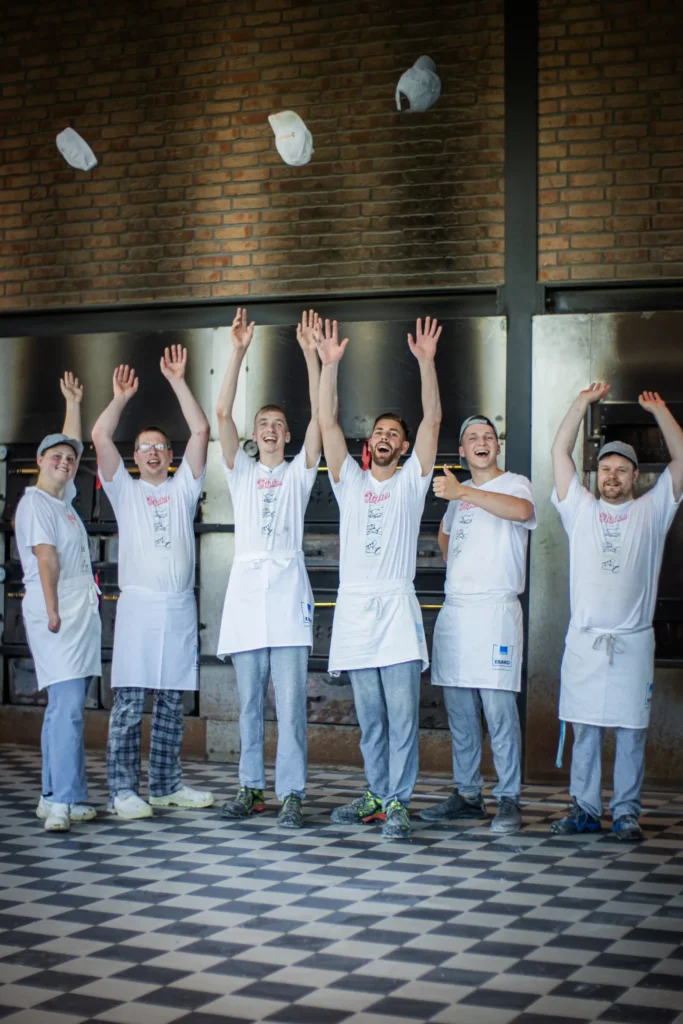 Sechs Bäcker in weißen Hemden und Schürzen stehen in einer Bäckerei, lächeln und heben ihre Arme, während sie ihre Hüte in die Luft werfen. Im Hintergrund sind Backsteinwände und Industrieöfen zu sehen.