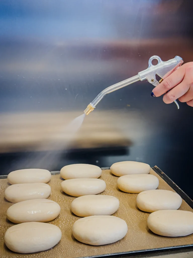 Eine Hand besprüht mit einer Sprühdüse ein Blech mit ungebackenen, ovalen Brötchen, die in Reihen auf einem Backblech angeordnet sind.