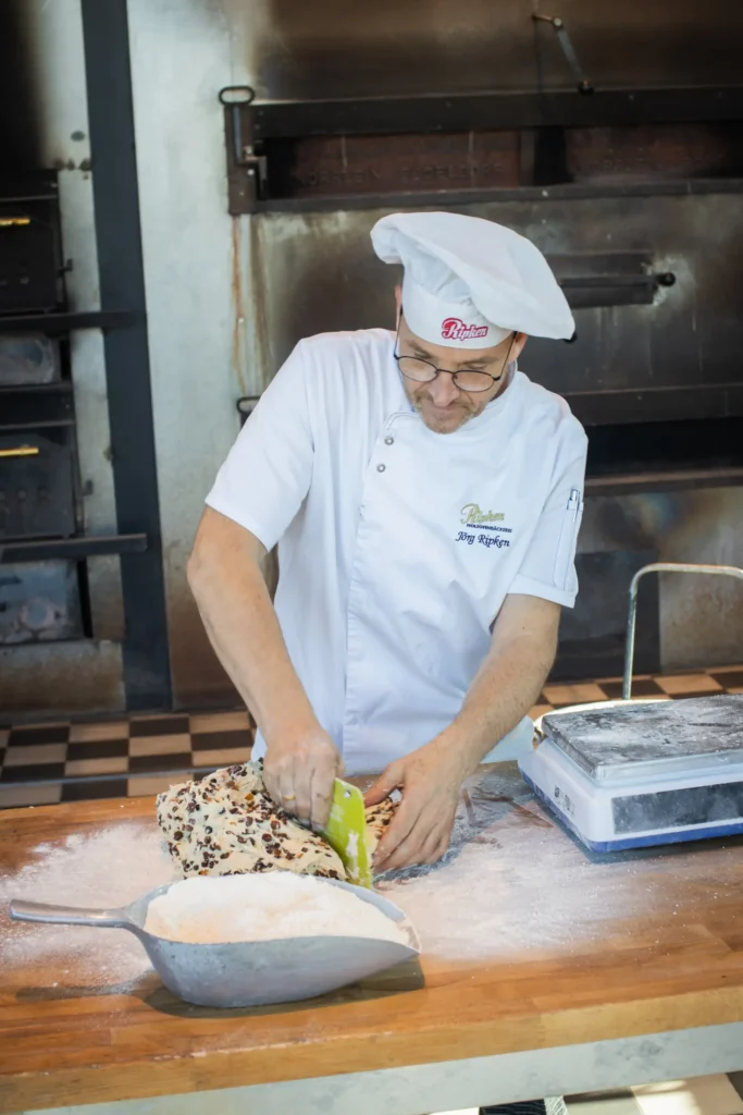 Ein Bäcker in weißer Uniform und mit Hut formt Teig mit Schokoladenstückchen auf einer bemehlten Fläche in einer Bäckereiküche, mit einem Industrieofen im Hintergrund.