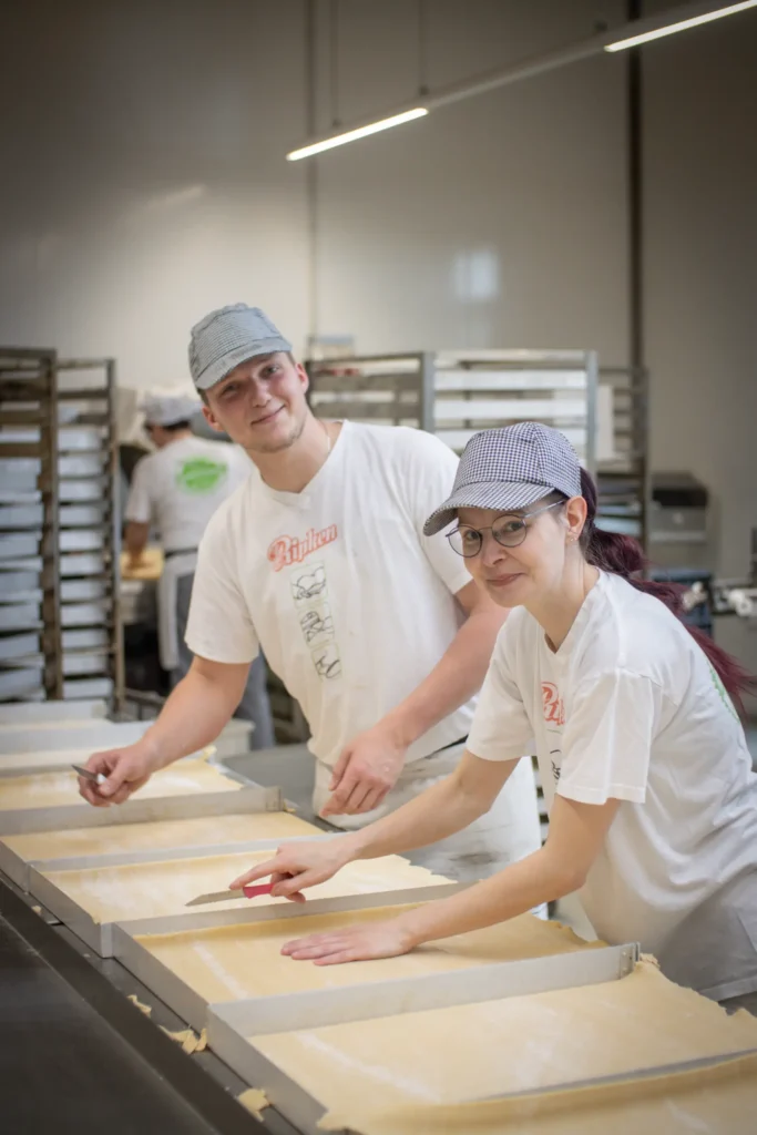 Zwei Personen mit Hüten und weißen Hemden arbeiten zusammen in einer Bäckerei und verteilen den Teig auf großen Blechen. Im Hintergrund sind Backständer und ein weiterer Arbeiter zu sehen. Beide Personen lächeln in die Kamera.