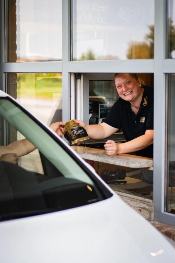Eine lächelnde Frau in schwarzer Uniform reicht einem Kunden durch ein Drive-Thru-Fenster eine Papiertüte. Die Hand des Kunden ist vom Inneren eines weißen Autos aus zu sehen.