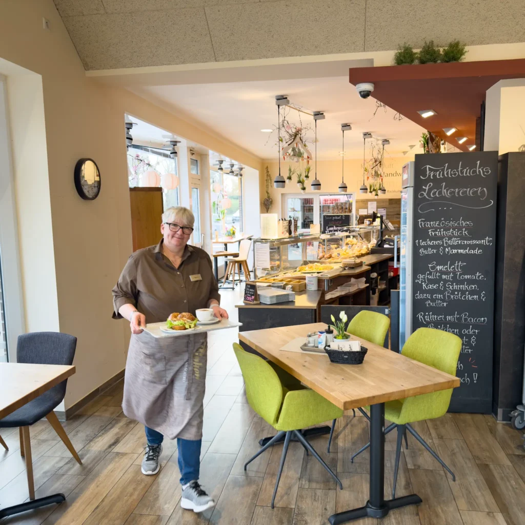 Eine lächelnde Bäckereifachverkäuferin trägt einen Teller mit Essen in ein gemütliches, modernes Café mit großen Fenstern, Holzböden, grünen Stühlen und einer Speisekarte auf einer Kreidetafel. Hinter dem Tresen stehen Gebäck und andere Lebensmittel.