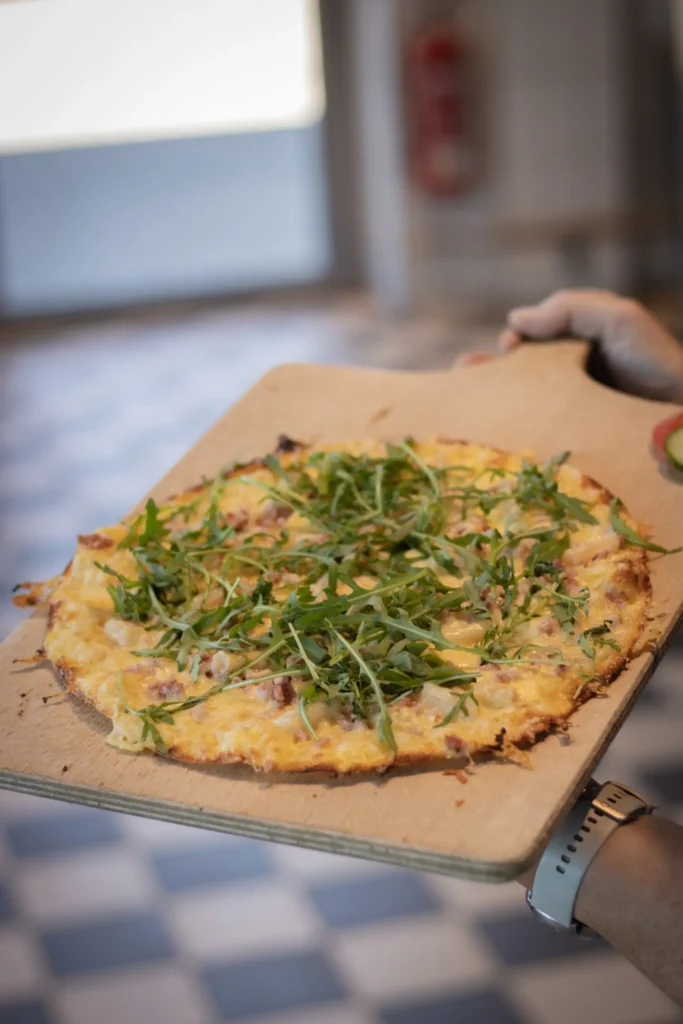 Eine Person hält ein Holzbrett mit einem Flammkuchen mit dünner Kruste, der mit Käse, Rucola und möglicherweise anderen Zutaten belegt ist. Der Hintergrund zeigt einen gefliesten Boden und eine unscharfe Innenumgebung.