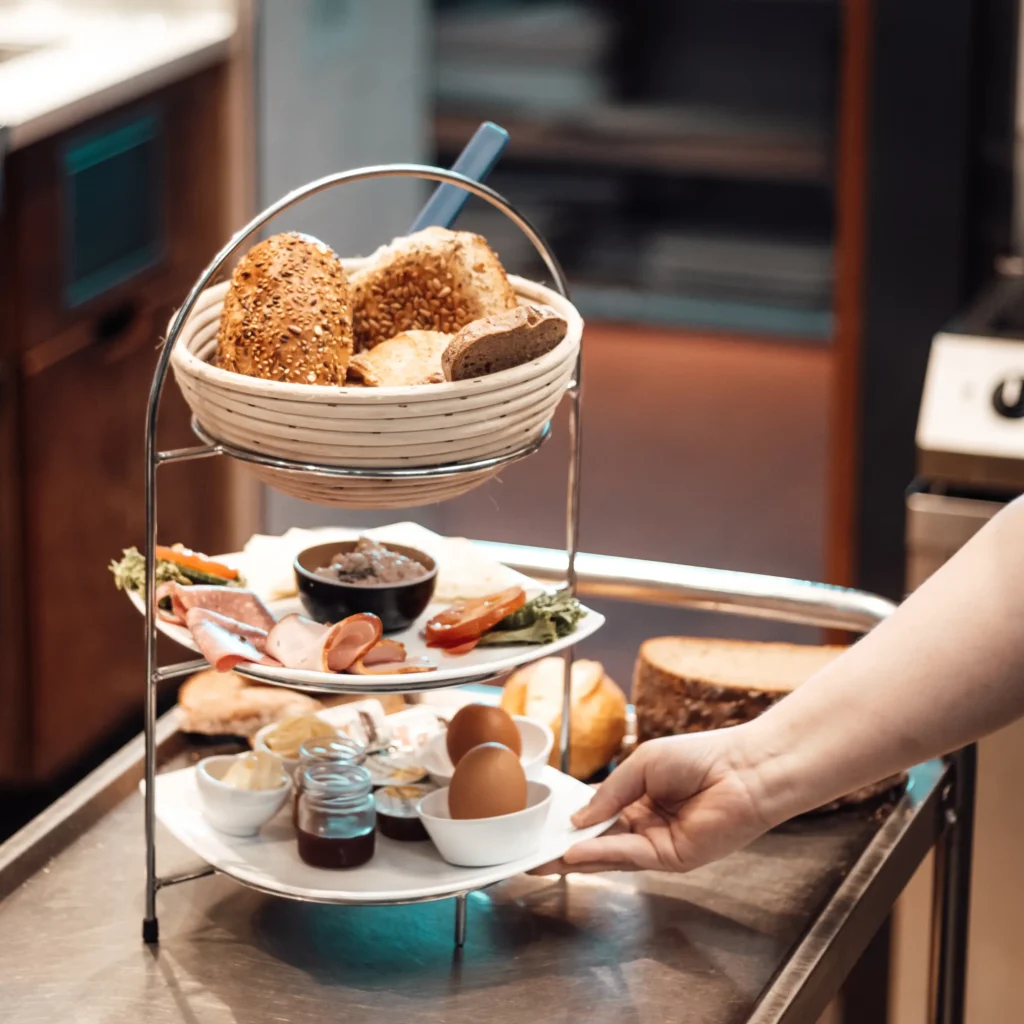 Eine Person hält eine zweistöckige Etagere mit verschiedenen Brotsorten auf der oberen Etage und verschiedenen Frühstücksprodukten, darunter gekochte Eier, Aufstriche, Aufschnitt und Marmelade aus dem Glas, auf der unteren Etage.