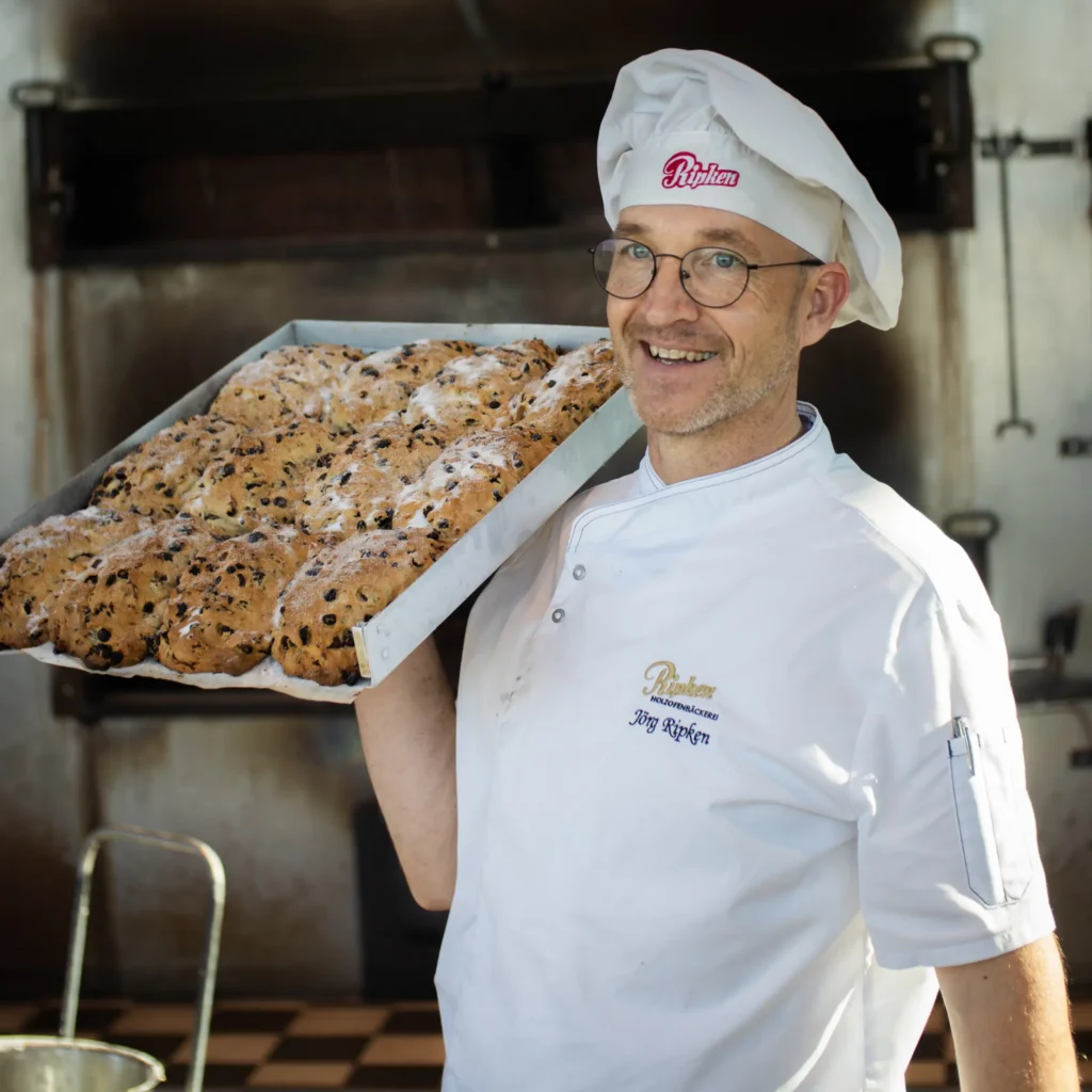 Ein lächelnder Bäcker mit Brille und weißer Uniform steht in einer Bäckereiküche und hält ein großes Tablett mit frisch gebackenem Brot, das mit Rosinen oder Schokoladenstückchen bestreut ist.