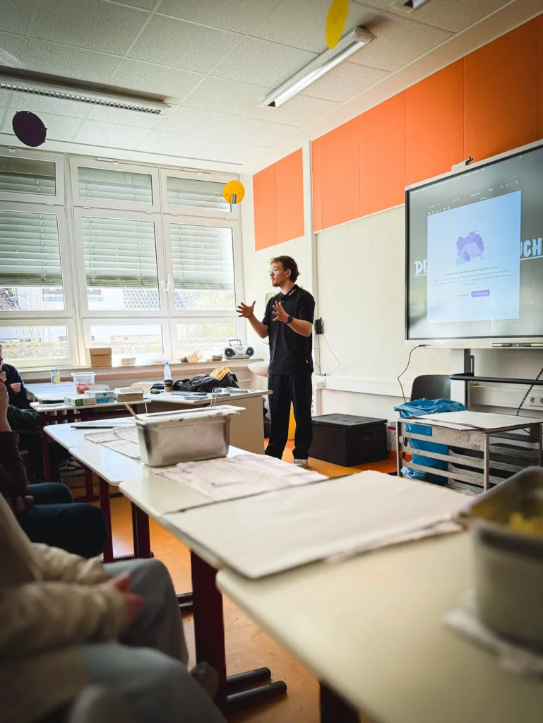 Eine Person steht und gestikuliert, während sie vor einer sitzenden Gruppe in einem Klassenzimmer eine Präsentation hält. Auf einem großen Monitor wird eine Website angezeigt, und auf den Tischen stehen Papiere und Behälter. Der Raum hat große Fenster und orangefarbene Wandpaneele.