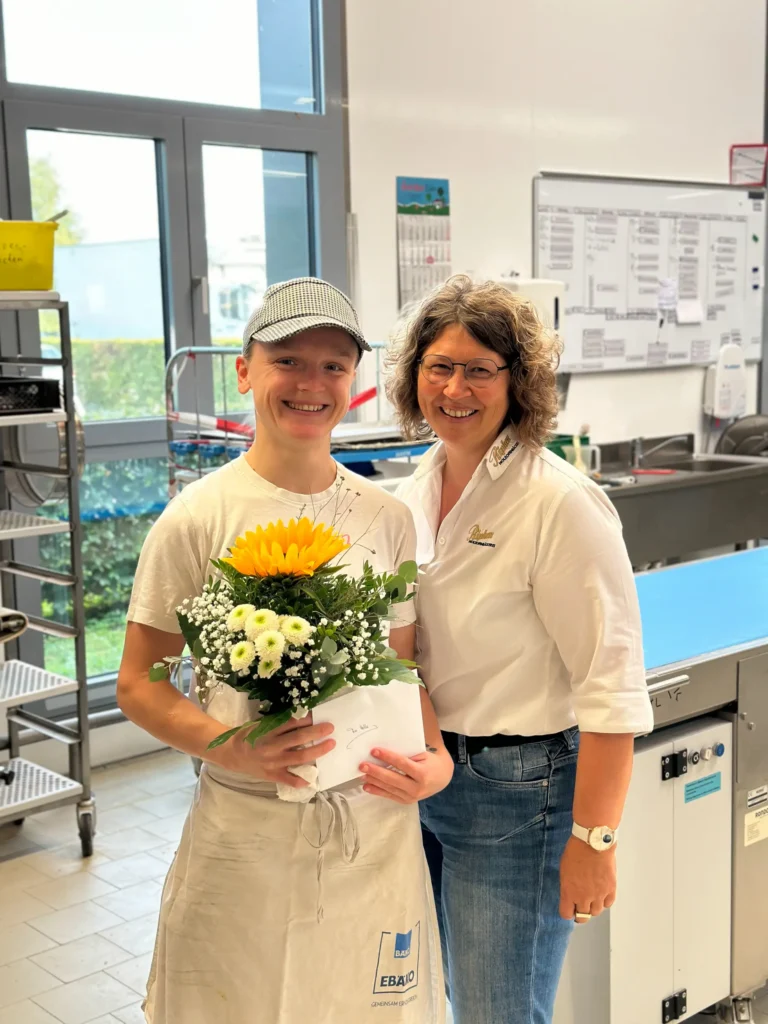 Zwei Frauen stehen lächelnd in einer hellen Küche. Die eine trägt eine Schürze und eine Mütze und hält einen Blumenstrauß und einen Umschlag in der Hand. Die andere, mit Brille und weißem Hemd, steht neben ihr. Hinter ihnen sind Küchengeräte und eine Zeittafel zu sehen.