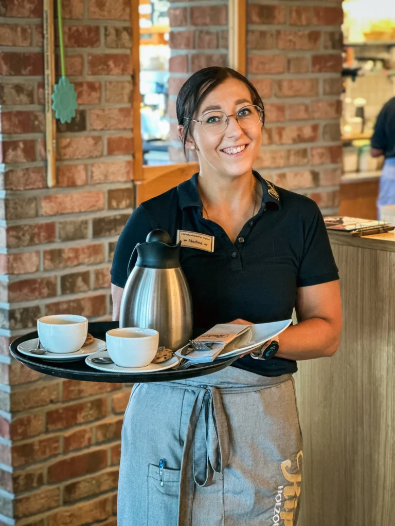 Eine lächelnde Kellnerin mit Brille und dunklem Polohemd hält ein Tablett mit zwei Kaffeetassen, Untertassen und einer metallenen Kaffeekanne in einem gemütlichen Café mit Backsteinwänden.