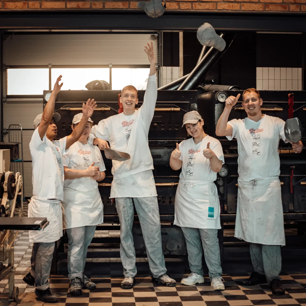 Fünf Bäcker in weißen Uniformen lächeln und posieren verspielt in einer Bäckerei und werfen Backwerkzeuge in die Luft. Sie stehen vor großen Öfen auf einem karierten Boden und wirken fröhlich und energiegeladen.