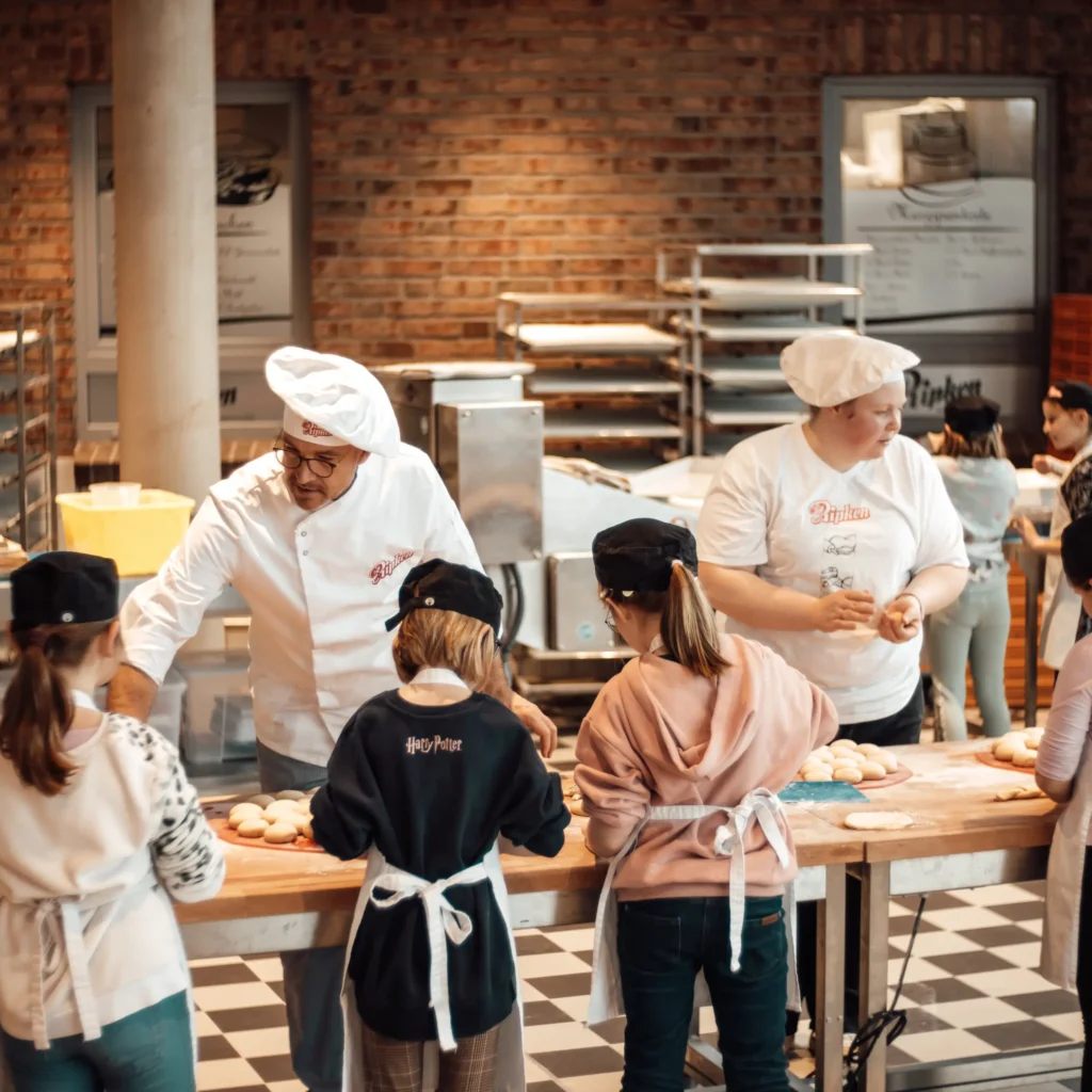 Eine Gruppe von Kindern mit Schürzen und Hüten versammelt sich um einen Bäckertisch und lernt von zwei erwachsenen Bäckern in einer hellen, industriellen Küche mit Regalen voller Bleche im Hintergrund, wie man Brotteig herstellt.