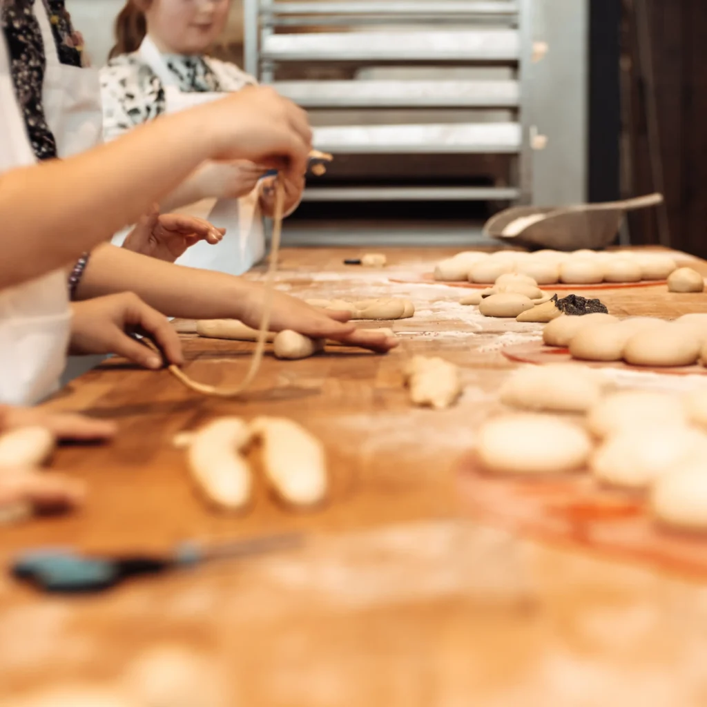 Kinder in Schürzen formen Teig auf einem bemehlten Holztisch, mit Backblechen und Küchenwerkzeugen im Hintergrund, was auf eine gemeinsame Back- oder Kochaktivität hindeutet.