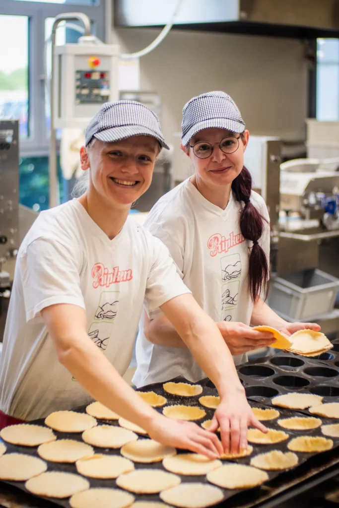 Zwei Personen mit weißen Hemden und karierten Mützen drücken lächelnd in einer Bäckereiküche Teig in Tortenformen. Um sie herum sind Backgeräte und mit Teig gefüllte Bleche zu sehen.