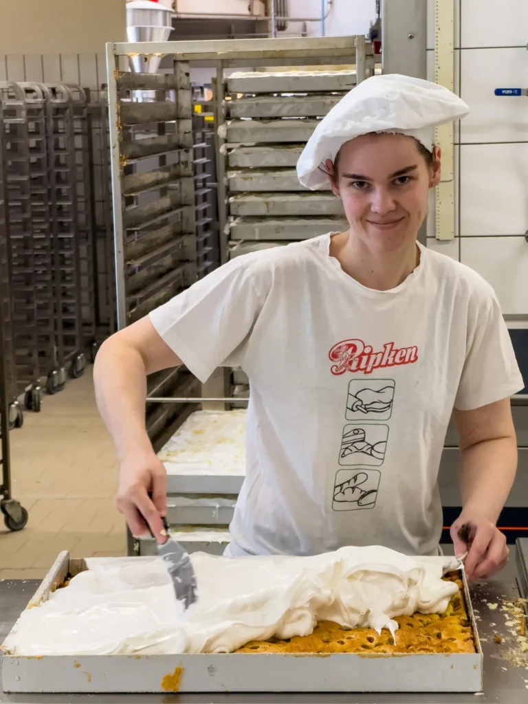 Eine Person in einer Bäckerei, die weiße Sahne auf ein großes Tablett mit Kuchen aufträgt. Sie trägt ein weißes Hemd und einen Papierhut und lächelt in die Kamera. Im Hintergrund sind hohe Metallgestelle zu sehen.