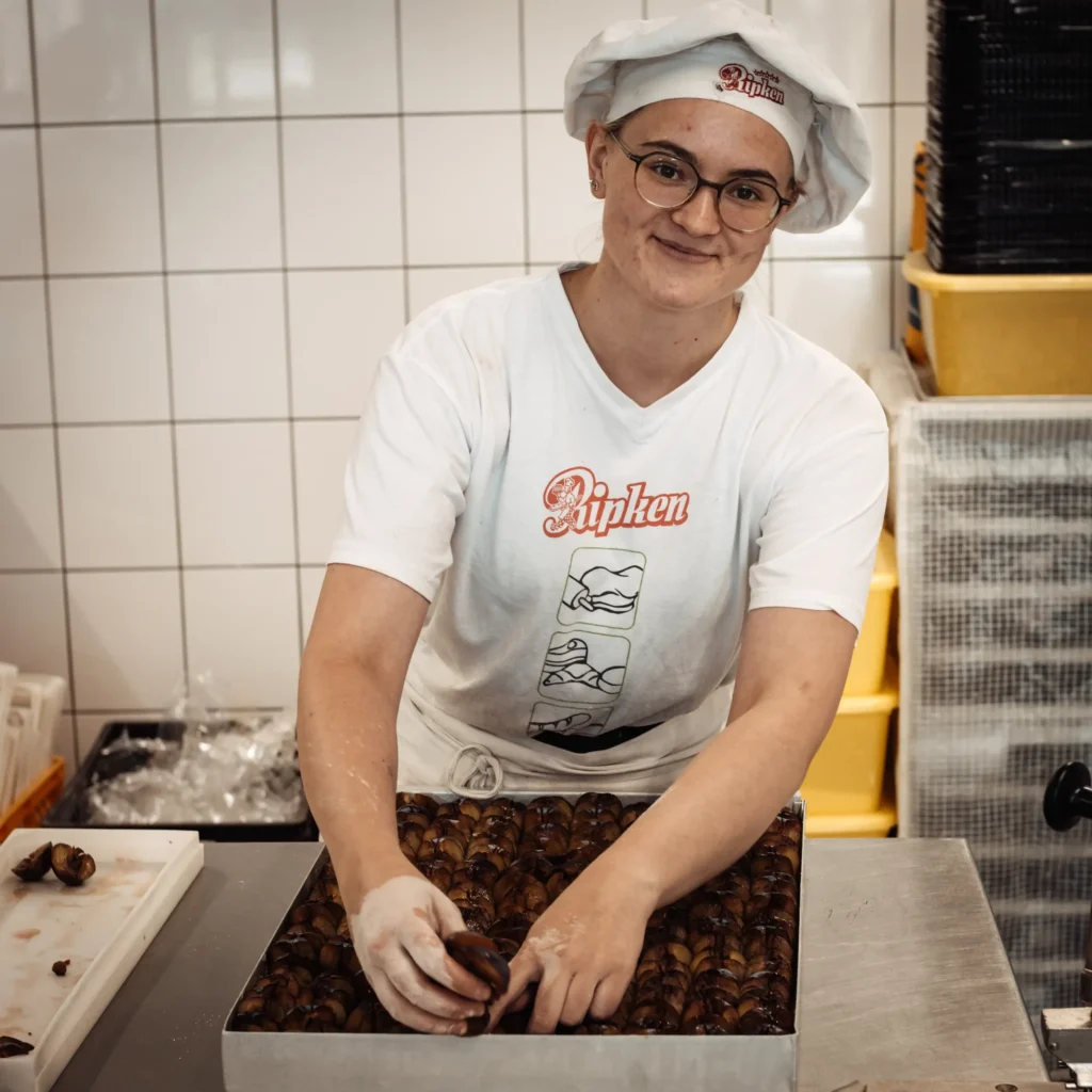 Eine Person, die ein weißes Hemd, eine Brille und eine Kochmütze trägt, ordnet lächelnd Gebäck auf einem Tablett in einer Bäckereiküche mit gekachelten Wänden und Backzubehör an.