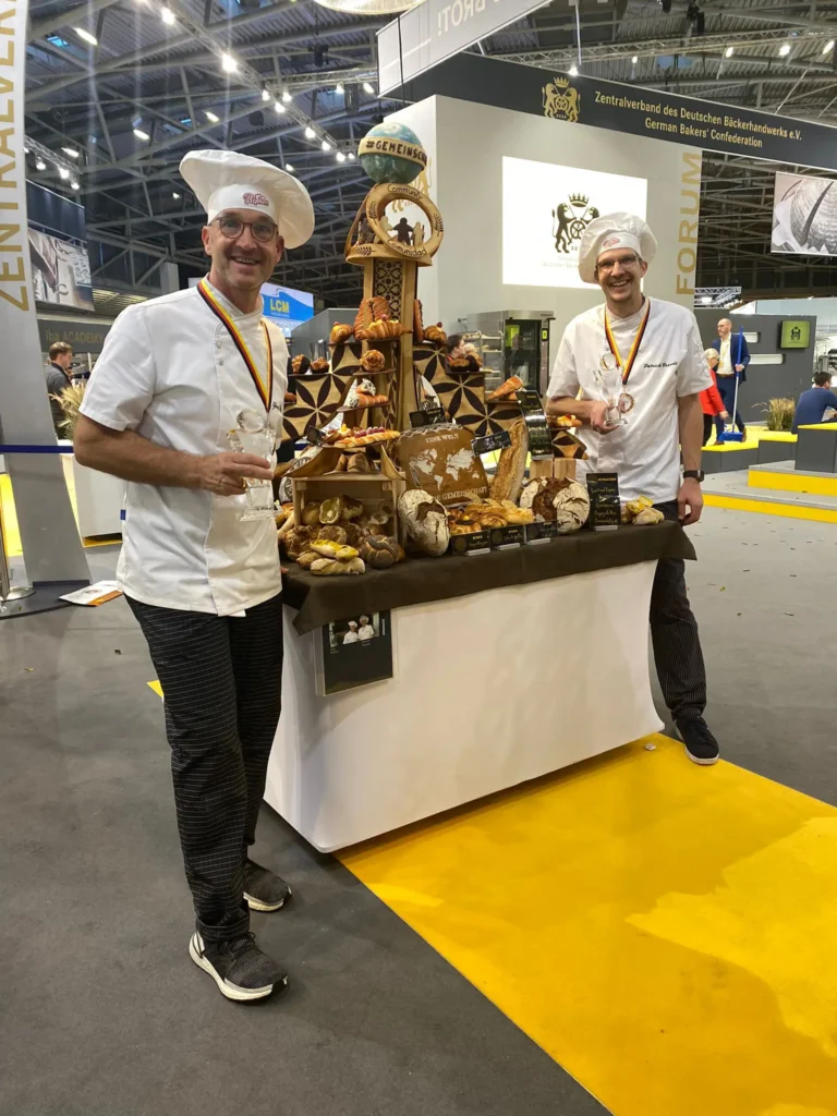 Zwei Bäcker mit weißen Kochmützen und Medaillen stehen lächelnd neben einem Ausstellungstisch mit verschiedenen Brotsorten und Gebäck auf einer Messe oder Ausstellung, während im Hintergrund andere Stände und Menschen zu sehen sind.