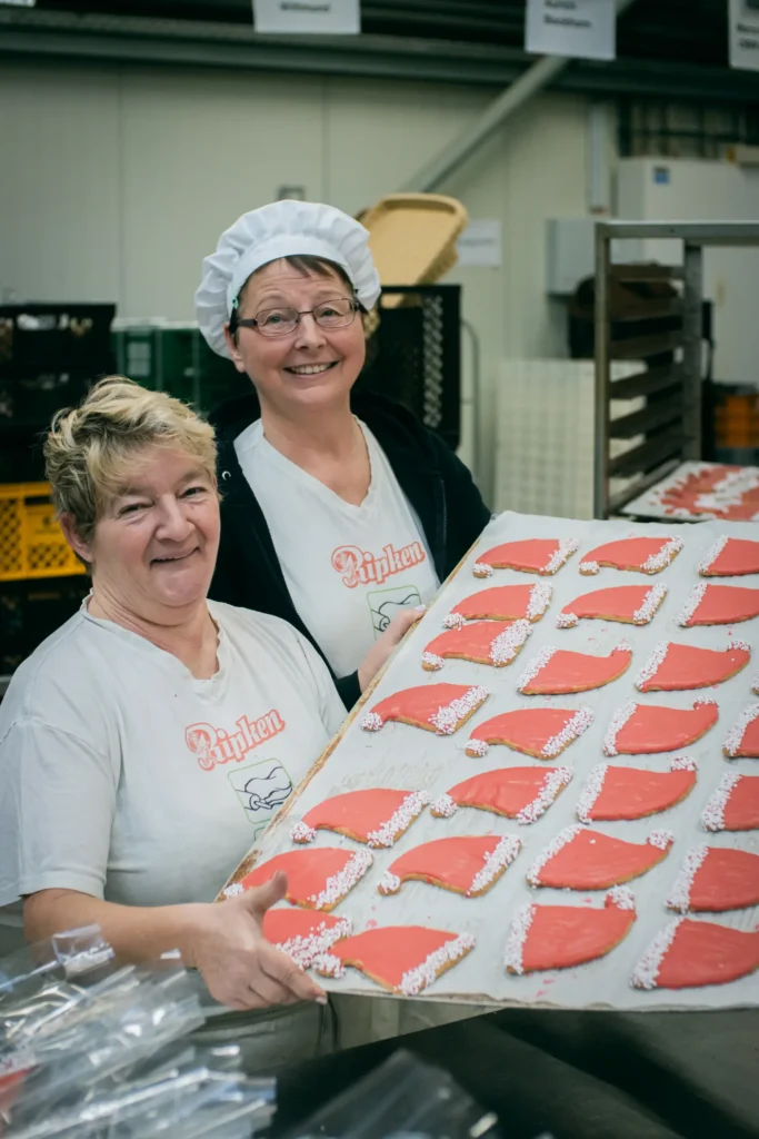 Zwei Frauen in weißen Hemden und Bäckerhüten lächeln, während sie in einer Großküche ein großes Tablett mit herzförmigen, rot glasierten Keksen mit weißem Zuckerguss in der Hand halten.