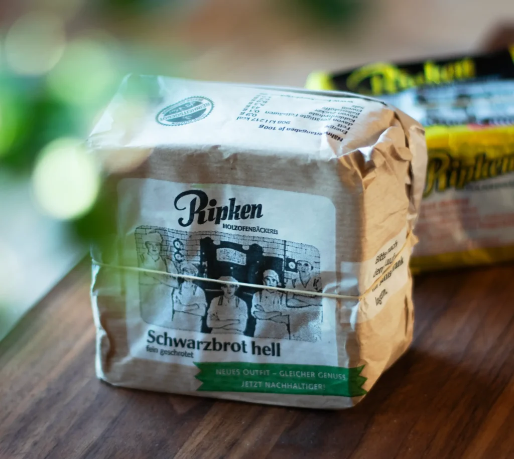 Eine braune Papierpackung des Schwarzbrotes hell liegt auf einer Holzfläche, mit grünen Blättern im Vordergrund und einem weiteren Laib Brot in gelber Verpackung dahinter.