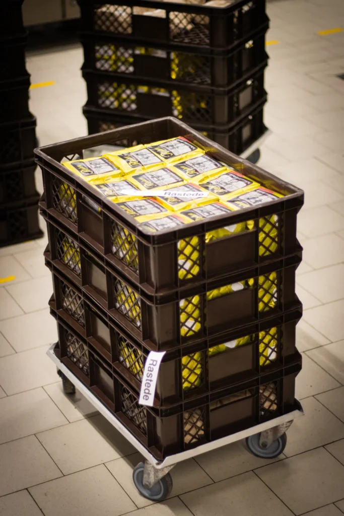 Ein Stapel brauner Kunststoffkisten, gefüllt mit gelber verpackter Ware, steht auf einem Rollwagen in einem gefliesten Innenbereich. An einer Seite der Kiste ist ein Etikett mit der Aufschrift "Rastedt" angebracht.