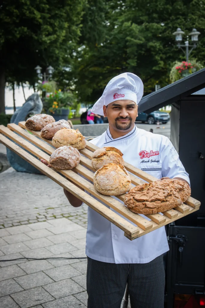 Ein lächelnder Bäcker in weißer Kochuniform und Mütze hält im Freien ein großes Holztablett mit mehreren frisch gebackenen Broten. Im Hintergrund sind Bäume, Blumen und ein Steinweg zu sehen.