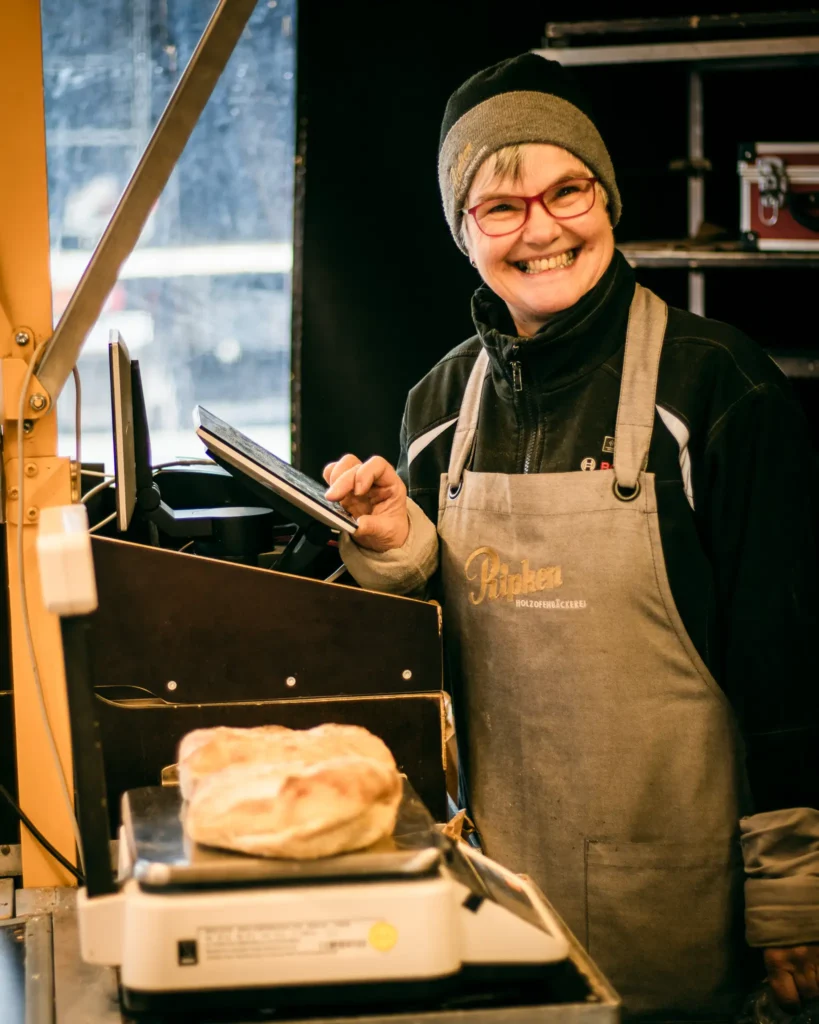 Eine lächelnde Person mit Brille, Mütze und Schürze steht an einem Tresen mit einer Digitalwaage und wiegt zwei Brote in einer Bäckerei.