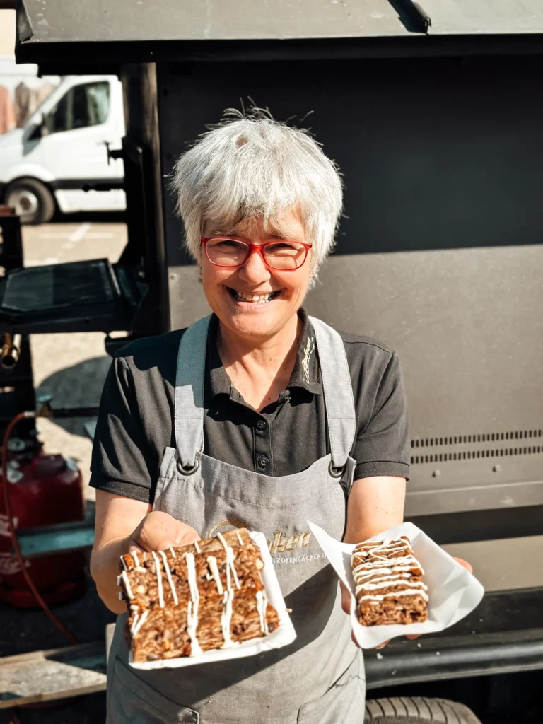 Eine lächelnde ältere Frau mit kurzen grauen Haaren und roter Brille hält zwei Stücke Kuchen oder Gebäck auf einem Teller und steht im Freien in der Nähe eines Imbisswagens oder -standes.