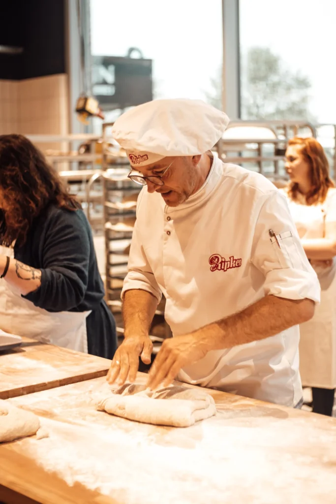 Ein Bäcker mit weißer Kochmütze und Kittel formt Teig auf einem bemehlten Tisch, während zwei weitere Personen in der Nähe in einer Bäckerei arbeiten.