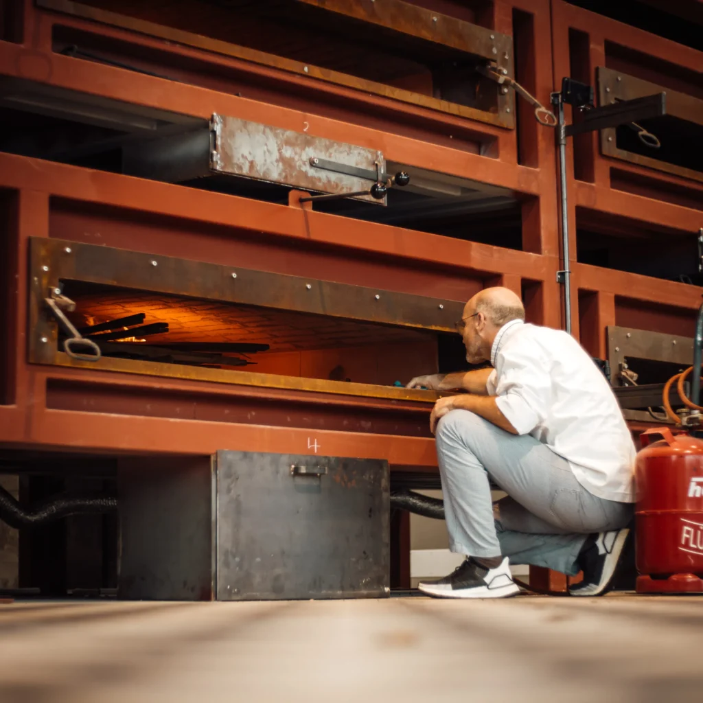 Ein Mann in weißem Hemd und grauer Hose hockt vor einem großen Industrieofen und blickt in eine beleuchtete, mit Ziegeln ausgekleidete Kammer. Der Ofen ist von roten Metallrahmen und Geräten umgeben.