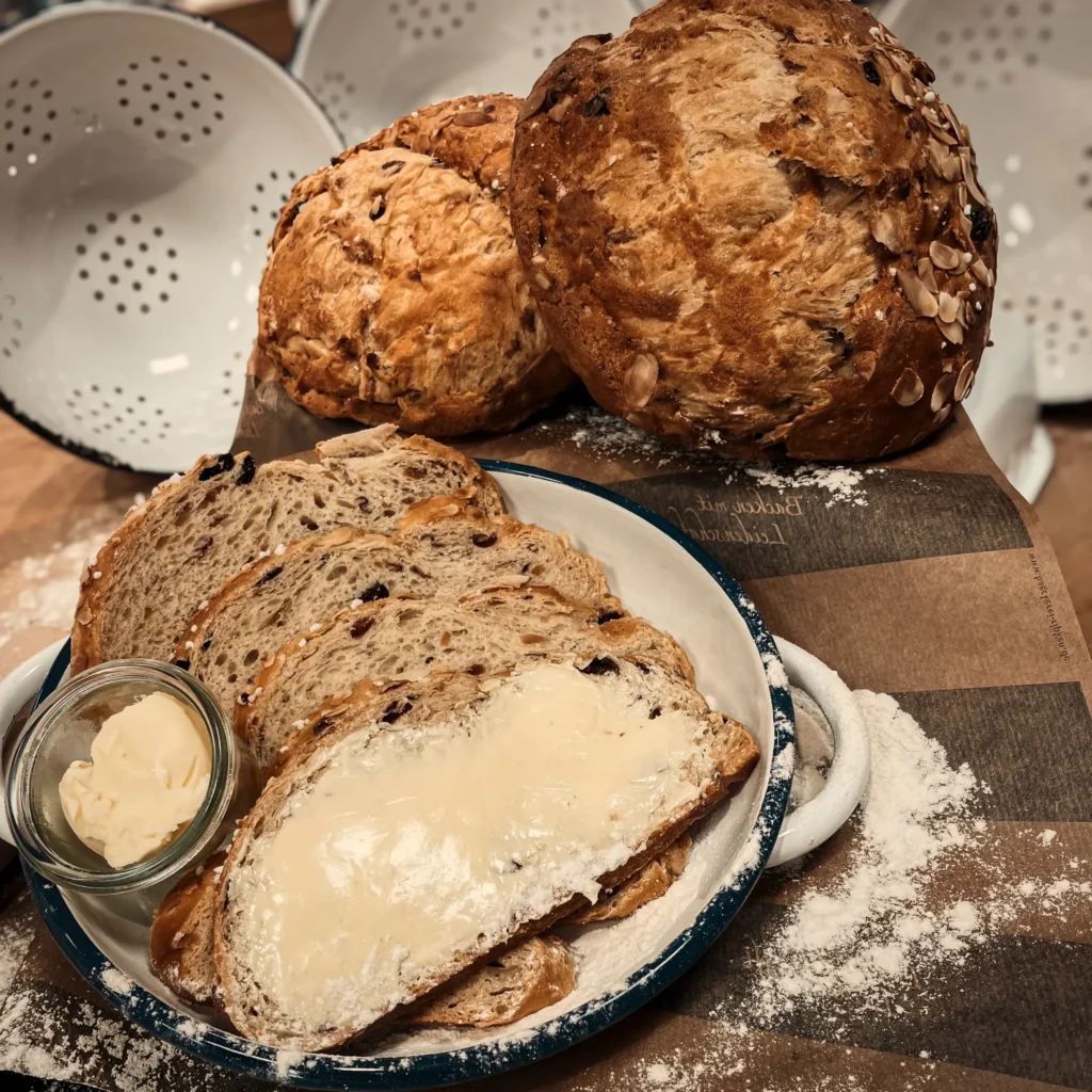 Ein Teller mit Brotscheiben, eine Scheibe mit Butter bestrichen, daneben eine kleine Glasschale mit Butter. Hinter dem Teller liegen zwei runde Laibe Bauernbrot. Die Holzfläche ist mit Mehl bestreut.