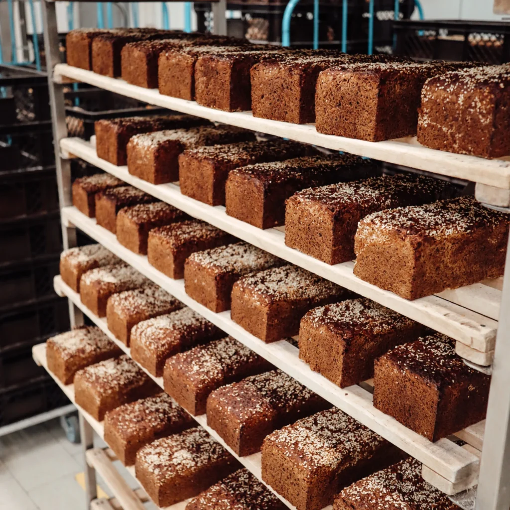 Reihen von rechteckigen, mit Körnern bedeckten Broten sind in einer Bäckerei fein säuberlich auf Holzgestellen angeordnet, bereit zum Abkühlen oder Verpacken. Im Hintergrund sind Metallregale und Bäckereimaschinen zu sehen.