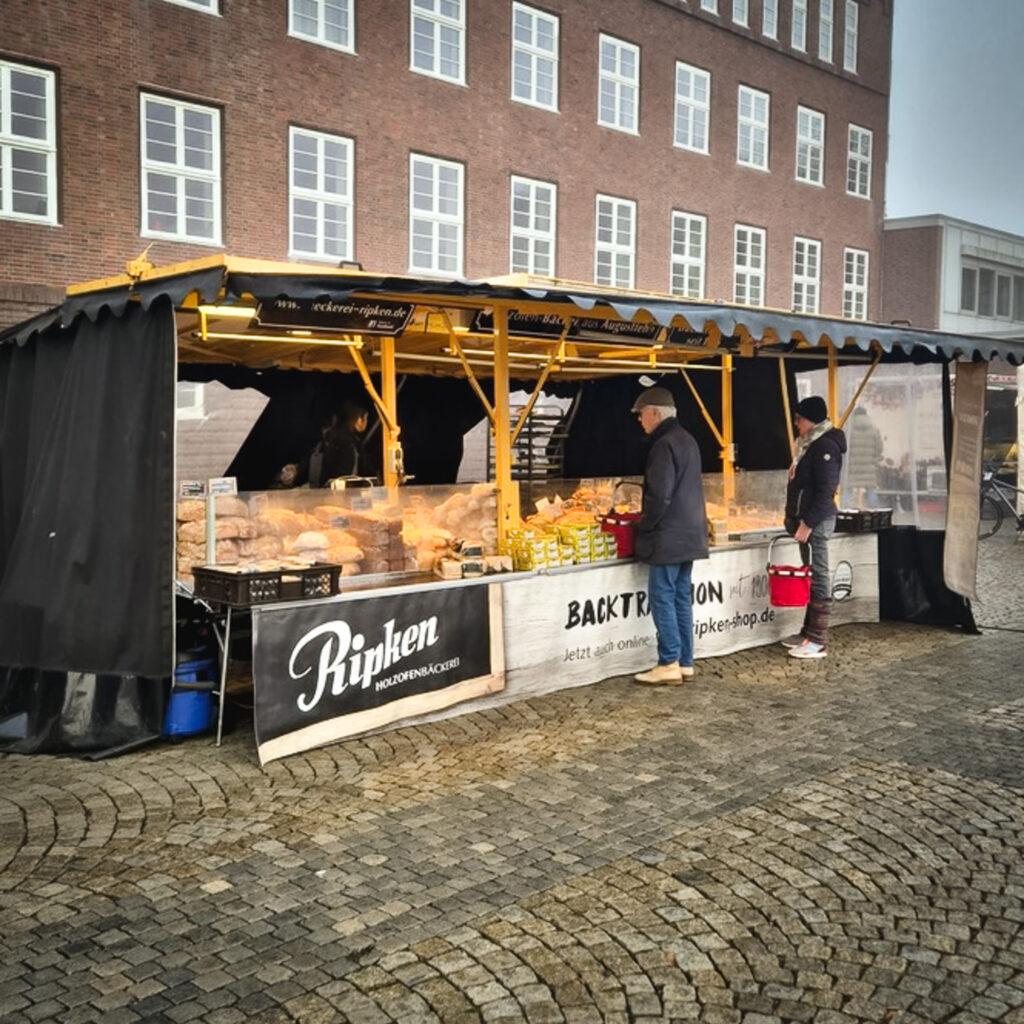 Ein Bäckereistand auf einem Markt im Freien bietet verschiedene Brotsorten und Backwaren an. Zwei Personen stehen davor und sehen sich das Angebot an. Der Stand hat ein schwarz-gelbes Vordach und befindet sich auf einem Platz mit Kopfsteinpflaster in der Nähe eines Backsteingebäudes.