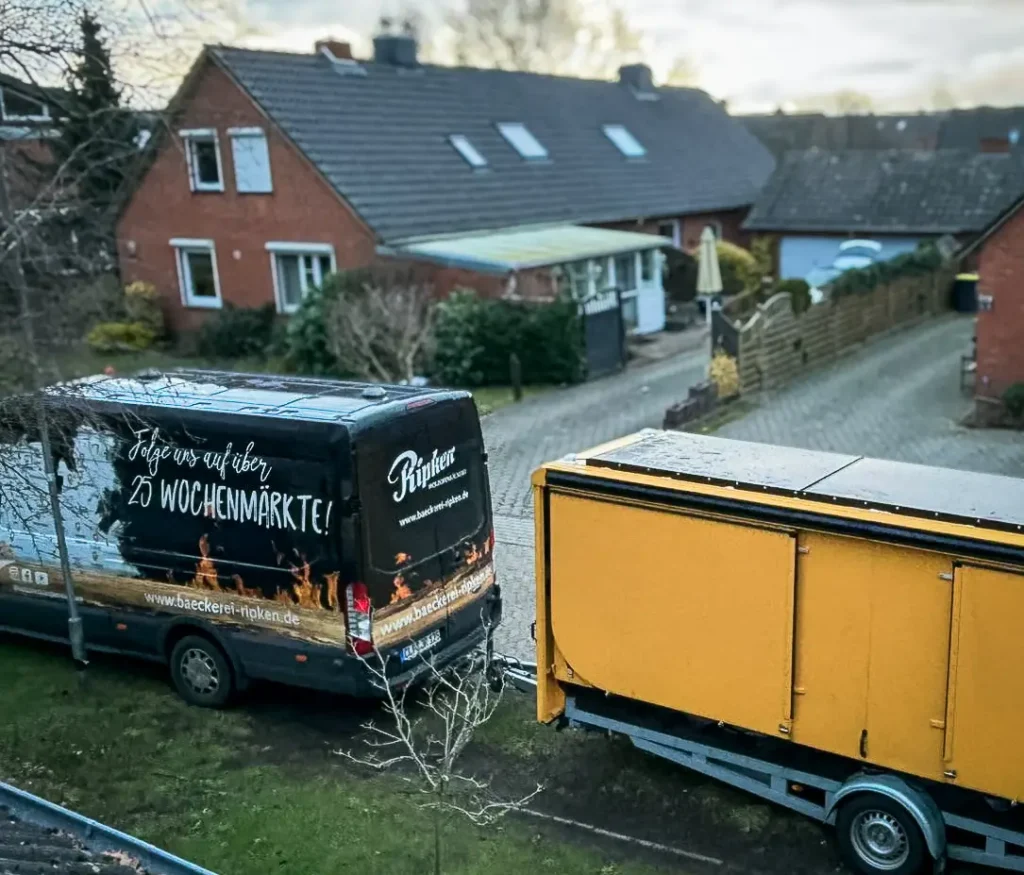 Zwei geparkte Lieferwagen in einer Wohngegend: ein schwarzer Lieferwagen mit deutscher Aufschrift, der für Wochenmärkte wirbt, und ein gelber Lieferwagen. Im Hintergrund sind Backsteinhäuser, Gärten und ein bewölkter Himmel zu sehen.