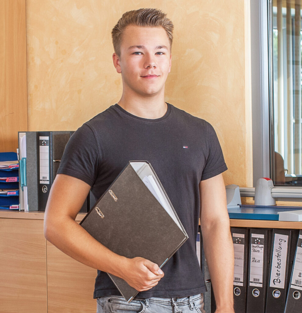 Ein junger Mann in einem schwarzen T-Shirt hält einen Aktenordner in der Hand und steht in einem Büro mit Regalen mit blauen Ordnern und Mappen, einem Fenster mit Jalousien und einem Holzschrank.