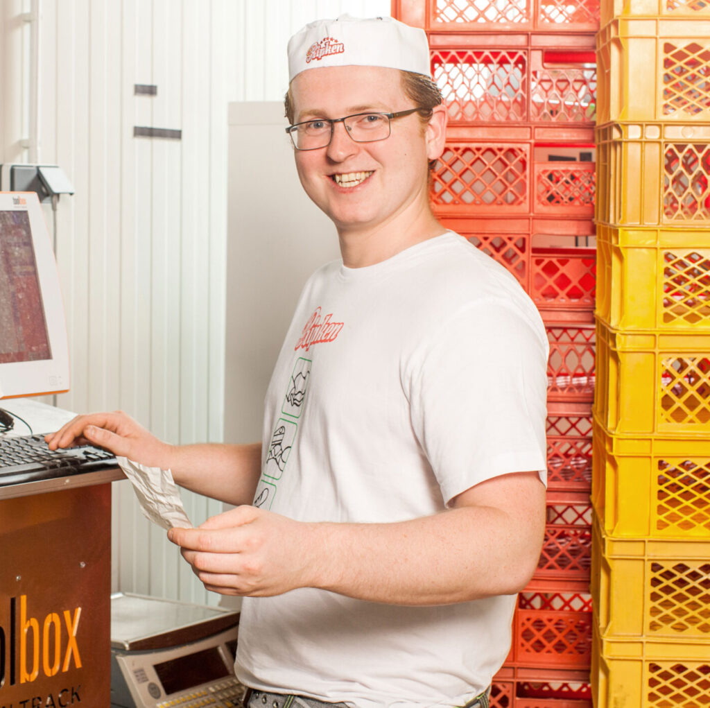 Ein lächelnder Bäcker mit weißer Mütze und weißem Hemd steht an einem Computer und hält einen Zettel in der Hand, daneben liegen Brotlaibe auf Regalen und bunte Plastikkisten in einer Bäckerei.