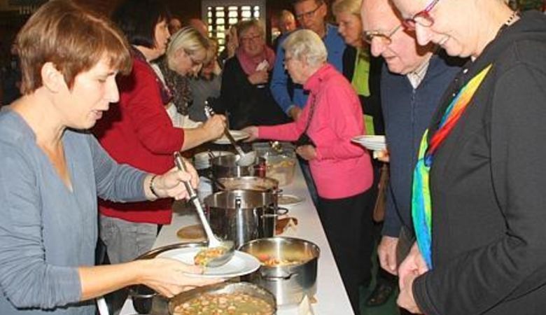 Eine Gruppe von Menschen steht in einer Schlange an einem Büffettisch und bekommt Suppe aus großen Töpfen serviert. Eine Frau im Vordergrund schöpft die Suppe in eine Schüssel, während andere mit Tellern warten.