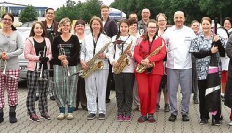 Eine Gruppe von Menschen, von denen einige Musikinstrumente wie Trompeten und Saxophone in der Hand halten, steht draußen auf einer gepflasterten Fläche. Viele von ihnen tragen Schlafanzüge und lächeln in die Kamera, was auf eine zwanglose oder themenbezogene Veranstaltung schließen lässt.