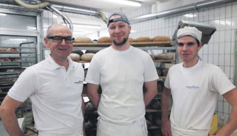 Drei Bäcker in weißen Uniformen und mit Hüten stehen lächelnd vor Regalen voller Brotlaibe in einer Bäckerei mit gekachelten Wänden und industriellen Geräten.