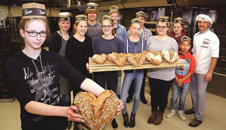Eine Gruppe von Menschen in einer Bäckerei, von denen viele Hüte tragen, posiert zusammen. Eine Person im Vordergrund hält einen herzförmigen Laib Brot, während andere weitere herzförmige Laibe auf Tabletts präsentieren. Ein Bäcker steht im Hintergrund und lächelt.
