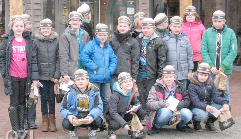 Eine Gruppe von Kindern in Wintermänteln und mit Papiermützen posiert für ein Foto im Freien. Sie halten kleine braune Tüten und Papierbögen in der Hand und stehen und knien vor einem Gebäude.