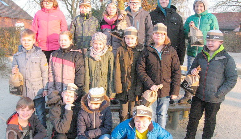 Eine Gruppe von Kindern in Winterkleidung und Mützen posiert an einem sonnigen Tag im Freien, einige sitzen auf einer Bank, andere stehen dahinter, halten kleine braune Taschen und lächeln in die Kamera.
