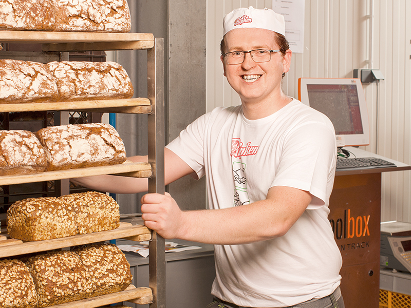 Ein lächelnder Bäcker mit Brille, weißem T-Shirt und Hut steht in einer Bäckerei neben einem Regal mit verschiedenen Brotsorten.