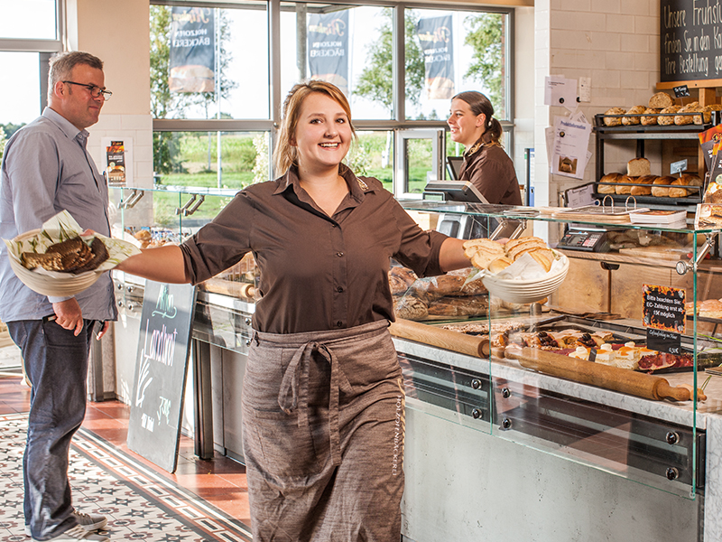 Eine lächelnde Frau in braunem Hemd und Schürze trägt Teller mit Essen in eine helle Bäckerei, in der ein Mann an der Theke wartet und eine andere Frau hinter der mit Backwaren gefüllten Glasauslage bedient.