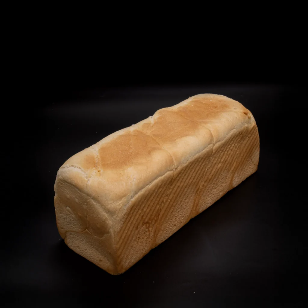 Ein rechteckiger Laib Weißbrot mit einer weichen, goldenen Kruste liegt auf einem schwarzen Hintergrund.