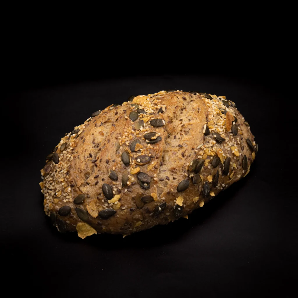 Ein rustikaler Laib Mehrkornbrot mit Samen, einschließlich Kürbis und Sesam, vor einem schwarzen Hintergrund.