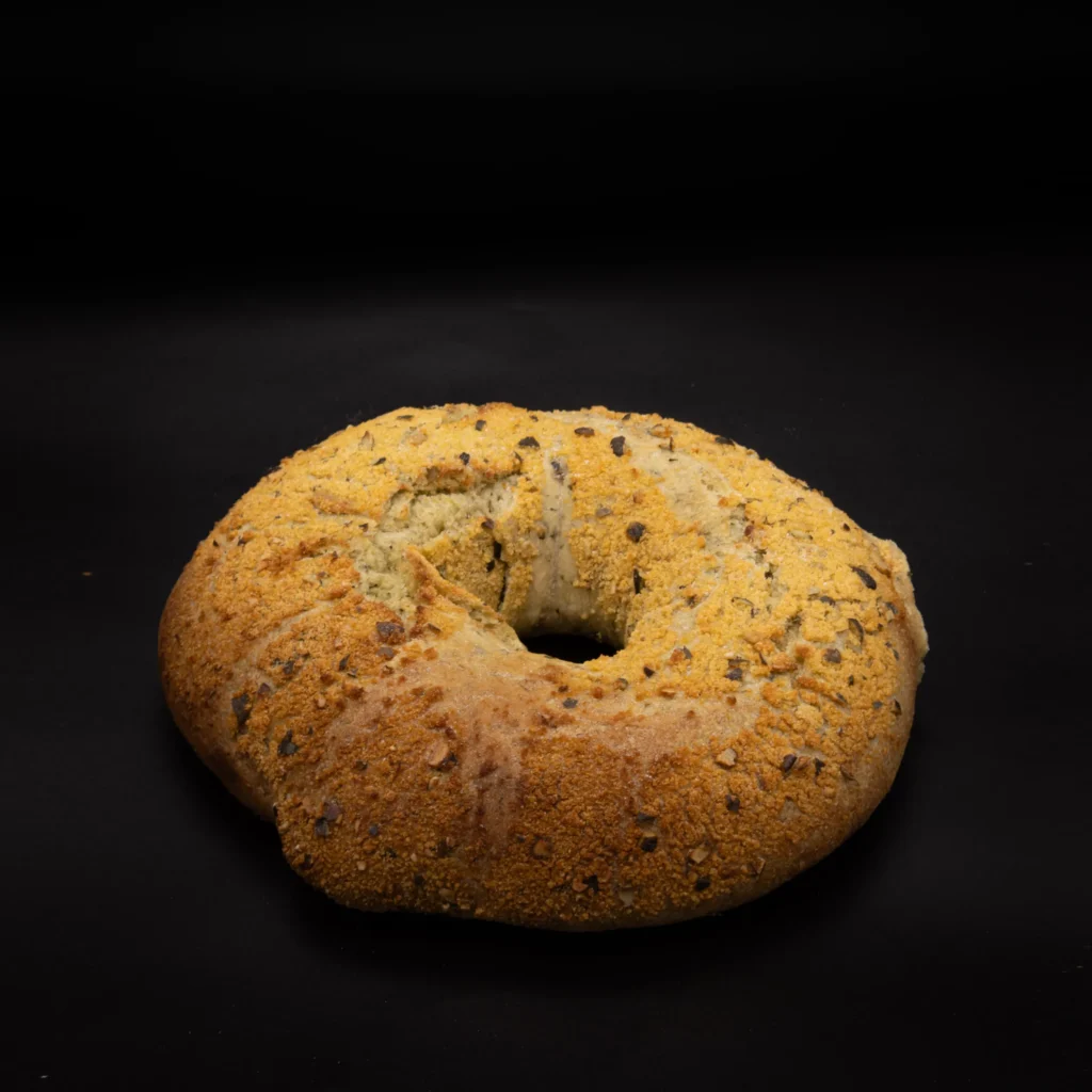Ein runder, rustikaler Brotlaib mit einem Loch in der Mitte, belegt mit einer goldenen, leicht knusprigen Kruste und verstreuten Samen, vor einem schwarzen Hintergrund.