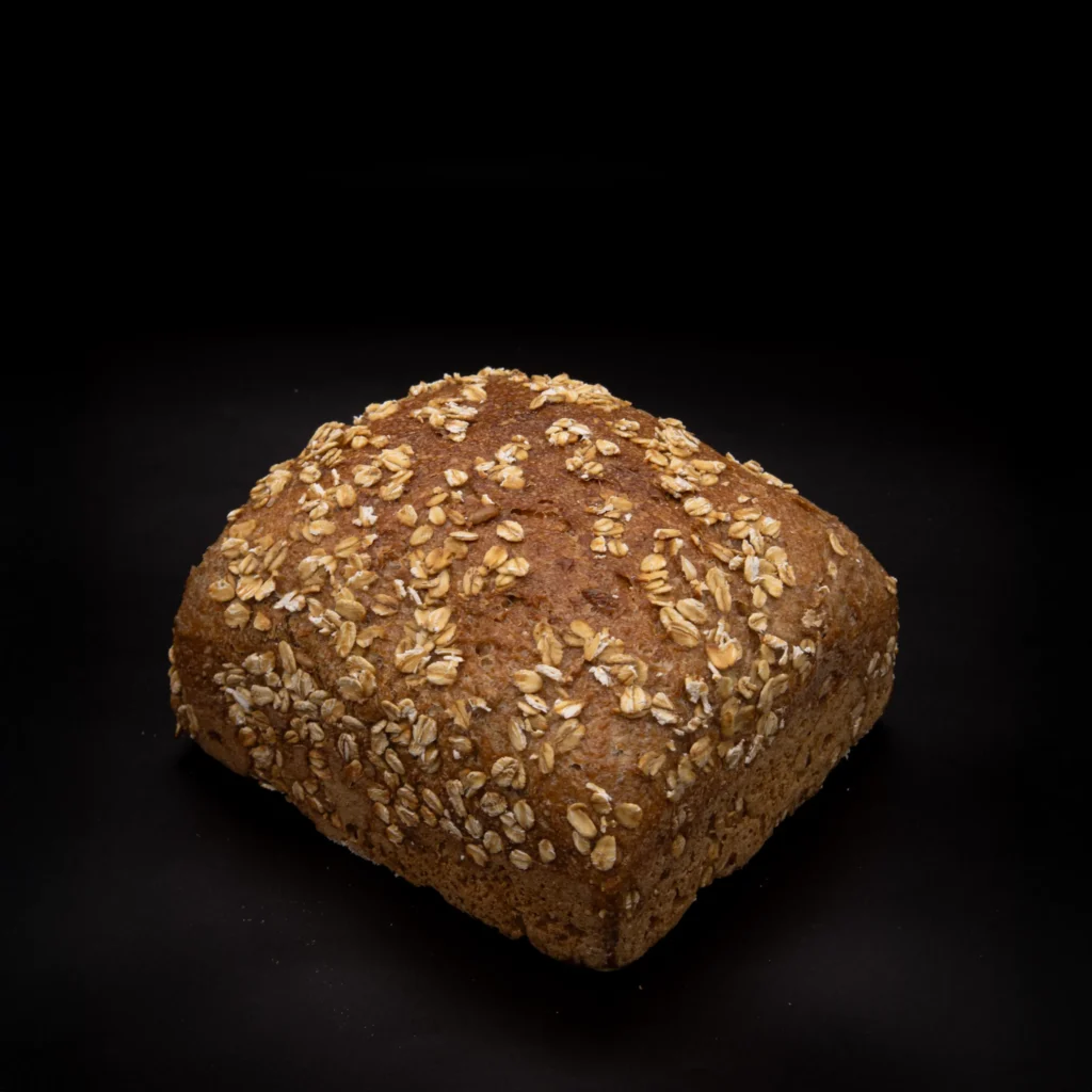 Ein quadratischer Laib Vollkornbrot mit Haferflocken liegt auf einem schwarzen Hintergrund.