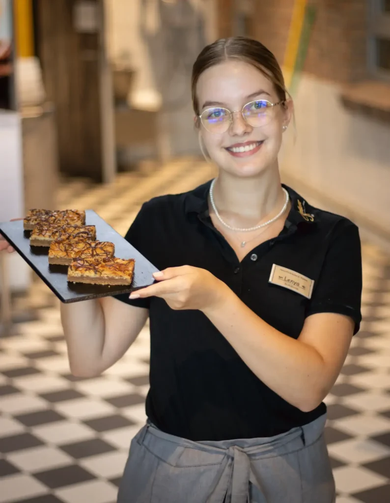 Eine lächelnde Frau im schwarzen Hemd und mit Brille hält ein rechteckiges Tablett mit vier Desserttafeln in einem Café mit kariertem Boden. Sie trägt ein Namensschild und eine Perlenkette.