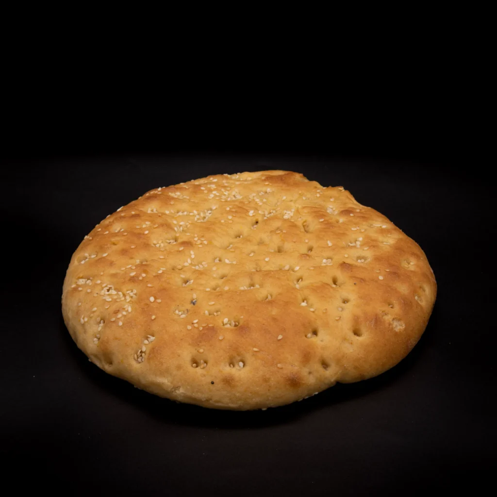 Ein rundes, goldenes Fladenbrot mit Sesam bestreut auf schwarzem Grund. Das Brot hat eine leicht gewellte Oberfläche und ein rustikales Aussehen.