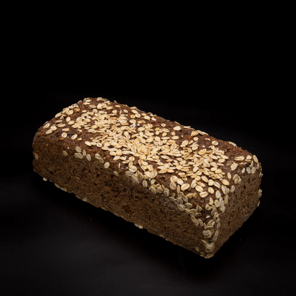 Ein rechteckiger Laib aus dunkelbraunem Brot mit Haferflocken, vor einem schlichten schwarzen Hintergrund.